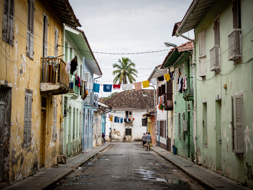 Wąska ulica w Centro Habana z wyblakłymi kolonialnymi kamienicami i przechodniami, palma w tle na tle nieba