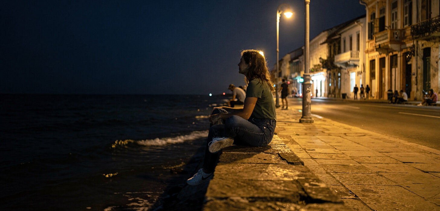 Kobieta siedząca na murku nabrzeża Malecón wieczorem, w tle oświetlone kolonialne budynki Vedado