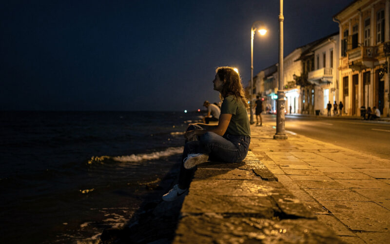 Kobieta siedząca na murku nabrzeża Malecón wieczorem, w tle oświetlone kolonialne budynki Vedado