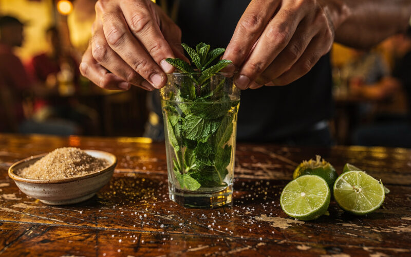 Ręce barmana delikatnie wyciskające świeże liście mięty do szklanki na drewnianym barze, obok kawałki limonki i cukier trzcinowy, w tle wnętrze małego kubańskiego baru z lokalnymi gośćmi