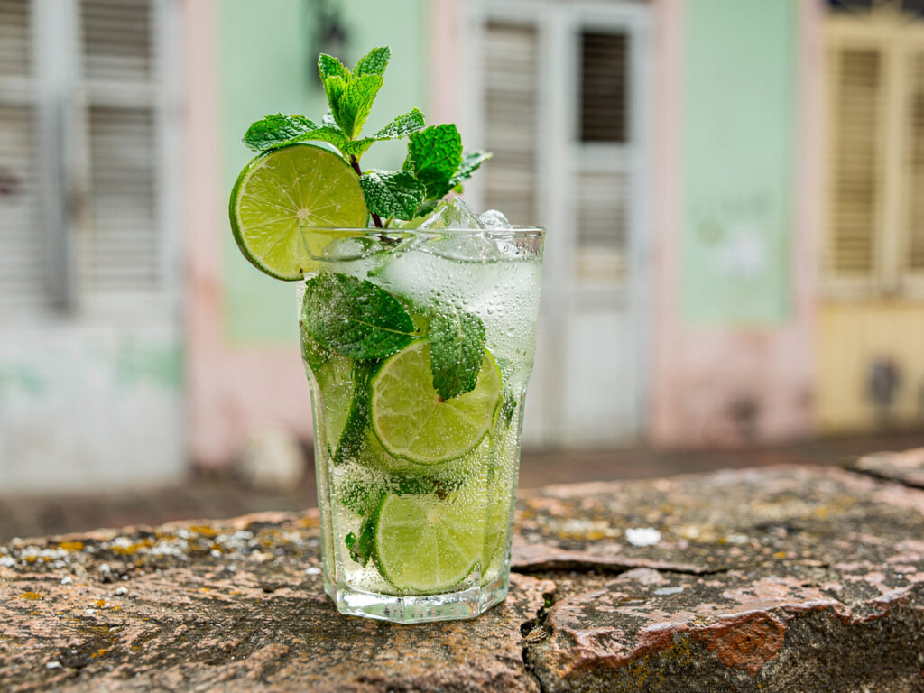 Szklanka mojito ze świeżą miętą i plasterkiem limonki na kamiennym murku, w tle rozmyta pastelowa fasada kolonialnej kamienicy w tropikalnym cieniu